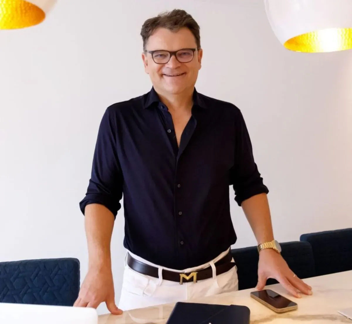 Christian Mugrauer mit Brille und dunklem Hemd, der Selbstbewusstsein ausstrahlt, steht mit einem Notebook und einem Smartpho