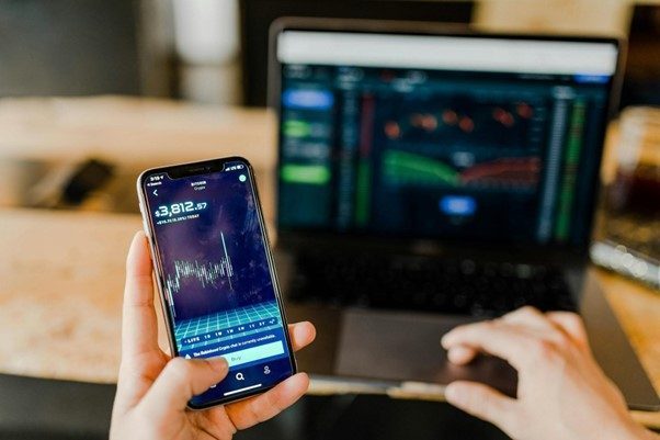 Person, die Daytrading betreibt, hält ein Smartphone mit einem Börsendiagramm in der Hand, im Hintergrund zeigt ein Laptop Finanzdaten an.