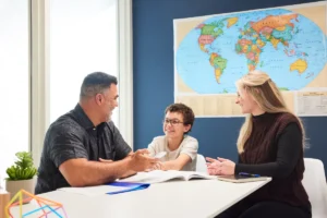 Drei Personen – zwei Erwachsene und ein Kind – sitzen mit offenen Büchern und Papieren an einem Tisch und diskutieren über Digitalisierung in Schulen. An der Wand hinter ihnen hängt eine Weltkarte, die an ein Klassenzimmer oder Büro mit Fokus auf moderner Bildung erinnert.