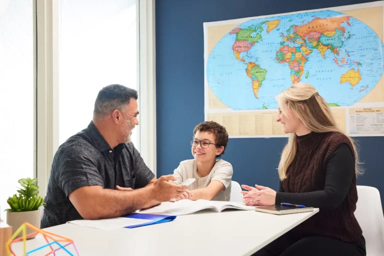 Drei Personen – zwei Erwachsene und ein Kind – sitzen mit offenen Büchern und Papieren an einem Tisch und diskutieren über Digitalisierung in Schulen. An der Wand hinter ihnen hängt eine Weltkarte, die an ein Klassenzimmer oder Büro mit Fokus auf moderner Bildung erinnert.