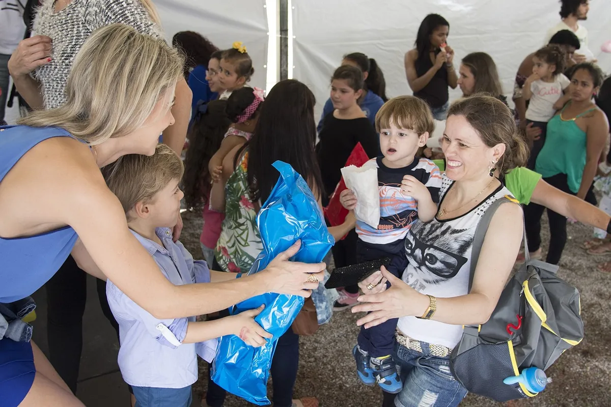 Eine Frau überreicht einem kleinen Kind ein blaues Paket. Eine andere Frau hält einen kleinen Jungen mit einem Rucksack. Im Hintergrund sind mehrere Kinder und Erwachsene zu sehen, die eine Atmosphäre schaffen, die eher an eine moderne Firmenweihnachtsfeier erinnert als an die üblichen langweiligen Feiertagsveranstaltungen.