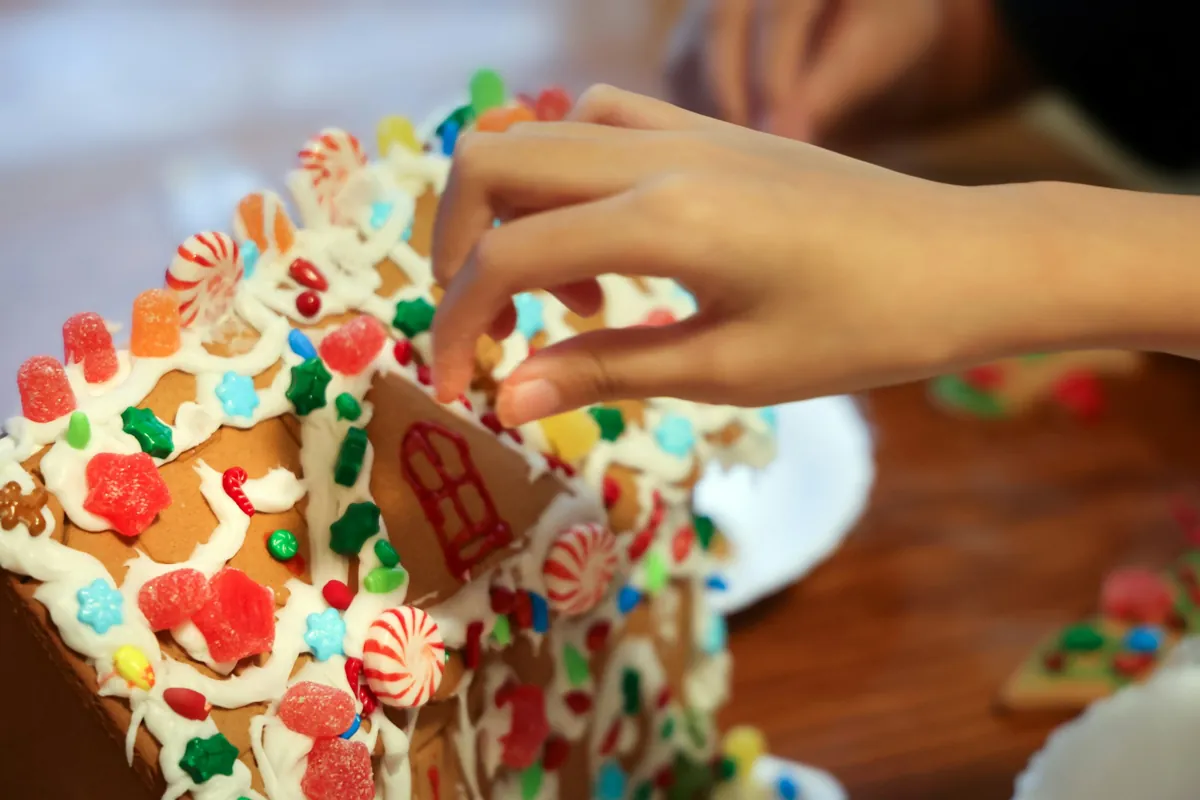 Eine Hand verziert ein Lebkuchenhaus mit bunten Süßigkeiten und Zuckerguss und verwandelt so eine alte Tradition in einen Schwall moderner Ideen, die sich perfekt für Firmenweihnachtsfeiern eignen.