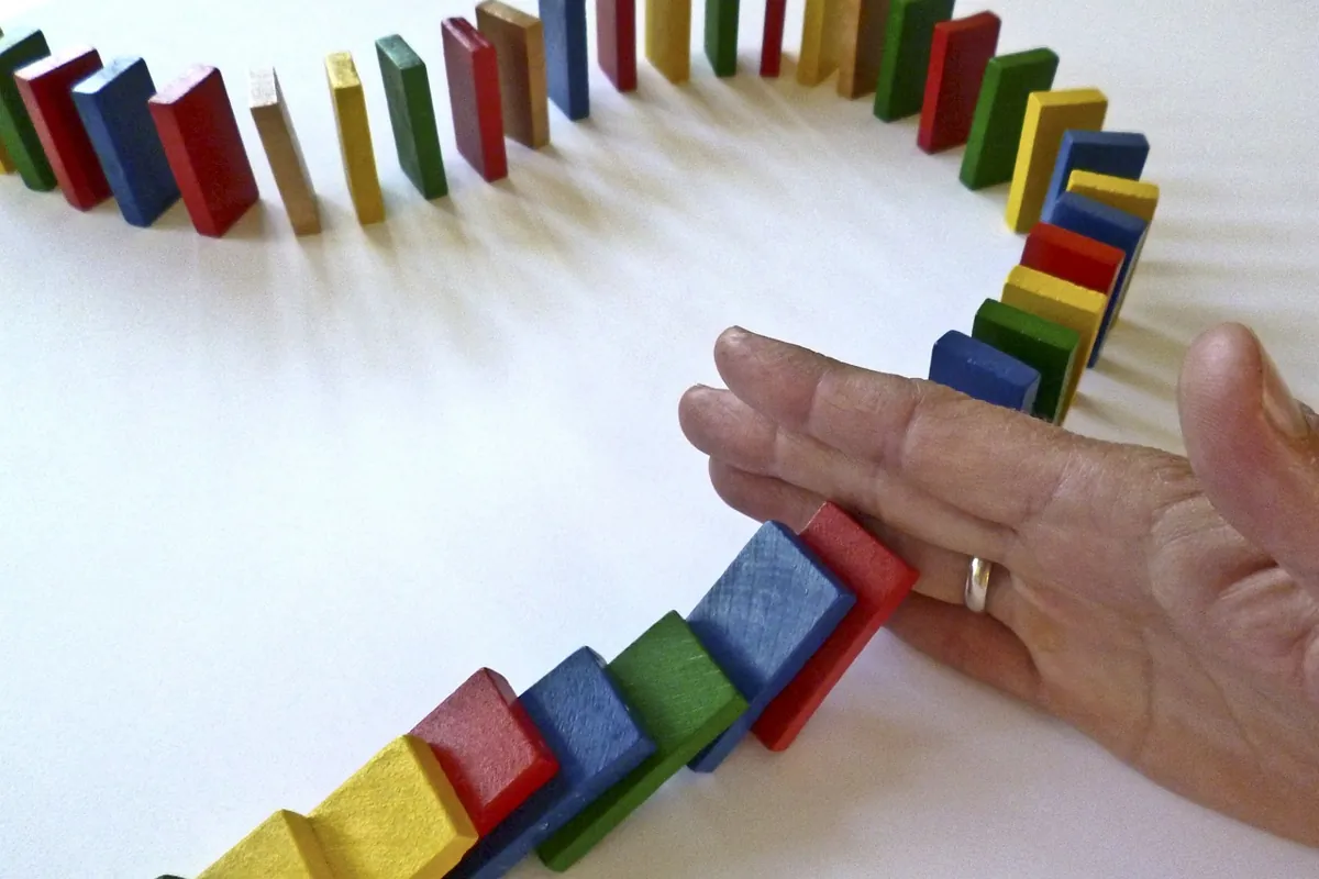 Eine Hand mit einem Ring am Finger stoppt eine Reihe bunter, fallender Dominosteine, die in einem Kreis auf einer weißen Fläche angeordnet sind, und symbolisiert den innovativen Ansatz, der erforderlich ist, um langweilige Firmenweihnachtsfeiern in unvergessliche Ereignisse zu verwandeln.