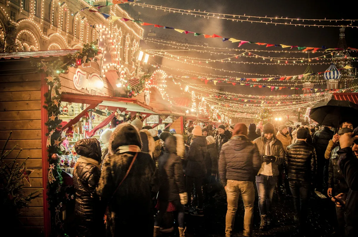 Ein geschäftiger Weihnachtsmarkt im Freien bei Nacht mit Menschen in warmer Kleidung, die zwischen dekorierten Ständen und festlichen Lichtern umherlaufen und moderne Ideen für Firmenweihnachtsfeiern bieten, damit die Feierlichkeiten alles andere als langweilig werden.