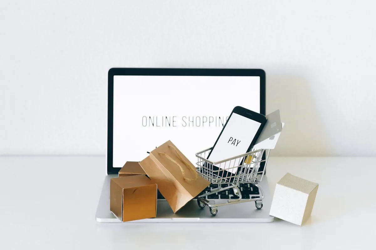 Auf einem Laptop steht der Schriftzug „Online Shopping“. Davor ist ein Miniatur-Einkaufswagen zu sehen, auf dem ein Smartphone mit dem Schriftzug „PAY“ und einer Kreditkarte steht, was die Essenz des E-Commerce einfängt. Daneben vervollständigen mehrere kleine braune und beige Einkaufstüten diese digitale Marktplatzszene.