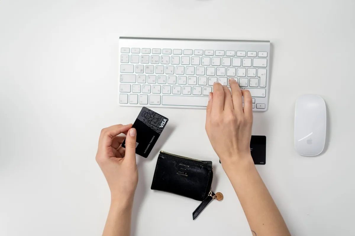 Eine Person verwendet eine weiße Tastatur und Maus auf einem weißen Schreibtisch und ist offensichtlich auf Online-Shopping konzentriert. In der linken Hand hält sie eine schwarze Kreditkarte und daneben eine schwarze Brieftasche. Sie ist bereit, das Beste aus ihrem E-Commerce-Erlebnis zu machen.