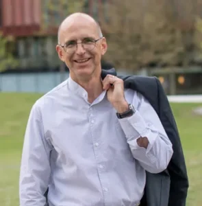 Peter Flume mit Brille und hellem Button-up-Hemd steht im Freien, lächelt in die Kamera und hält eine dunkle Jacke über die Schulter. Der erfahrene Rhetoriktrainer ist vor einem unscharfen Gebäude und Gras im Hintergrund zu sehen.