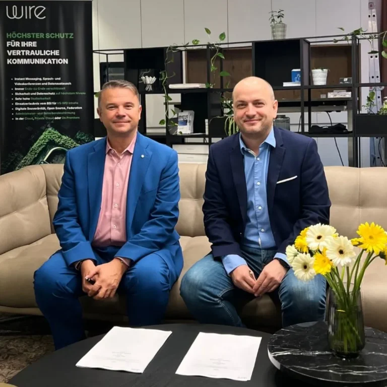 Zwei Männer in Geschäftskleidung sitzen auf einem beigen Sofa, auf dem Tisch liegen Papiere mit der Aufschrift "agilimo". Eine Vase mit gelben und weißen Blumen ziert die Szene, während Büroregale und ein stehendes Banner im Hintergrund eine strategische Partnerschaft andeuten.