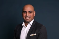 Firat Koeker in dunkler Anzugsjacke, weißem Hemd und Einstecktuch steht vor einem schlichten, dunklen Hintergrund und lächelt in die Kamera. Er verkörpert das Selbstvertrauen und die Innovationskraft der heutigen Green-Tech-Führungskräfte.