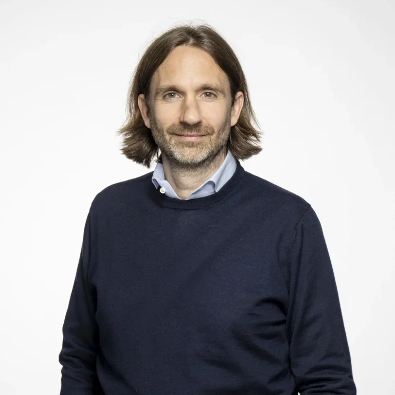 Michael Plankensteiner mit schulterlangen braunen Haaren und Bart steht vor einem schlichten weißen Hintergrund. Er trägt einen marineblauen Pullover über einem hellblauen Kragenhemd und verkörpert damit den professionellen Stil von Neuron Automation.