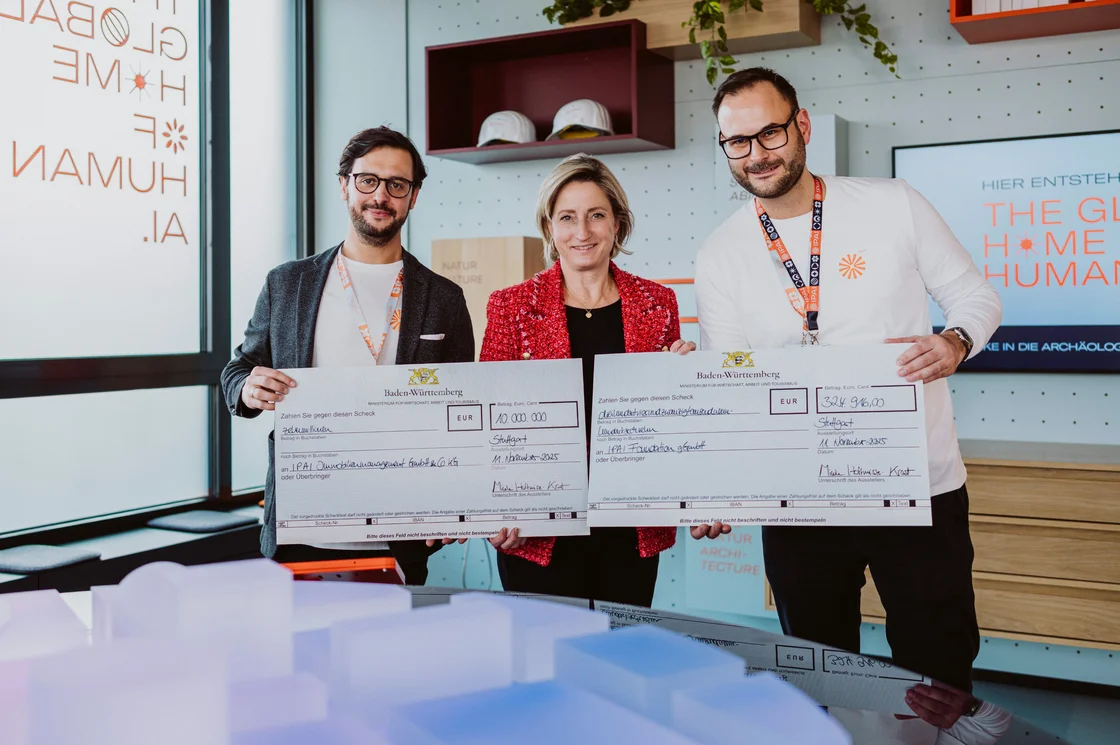 Drei Personen stehen in einem Innenraum und halten zwei große Präsentationsschecks für IPAI Förderung. Die lächelnden Personen blicken in die Kamera, während im Hintergrund ein Fenster mit Text und ein mit Büchern und Pflanzen bestücktes Regal zu sehen sind.